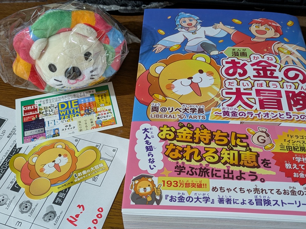alt="リベ大フェス2025で購入した書籍『漫画 お金の大冒険』とぬいぐるみキーホルダーの写真"