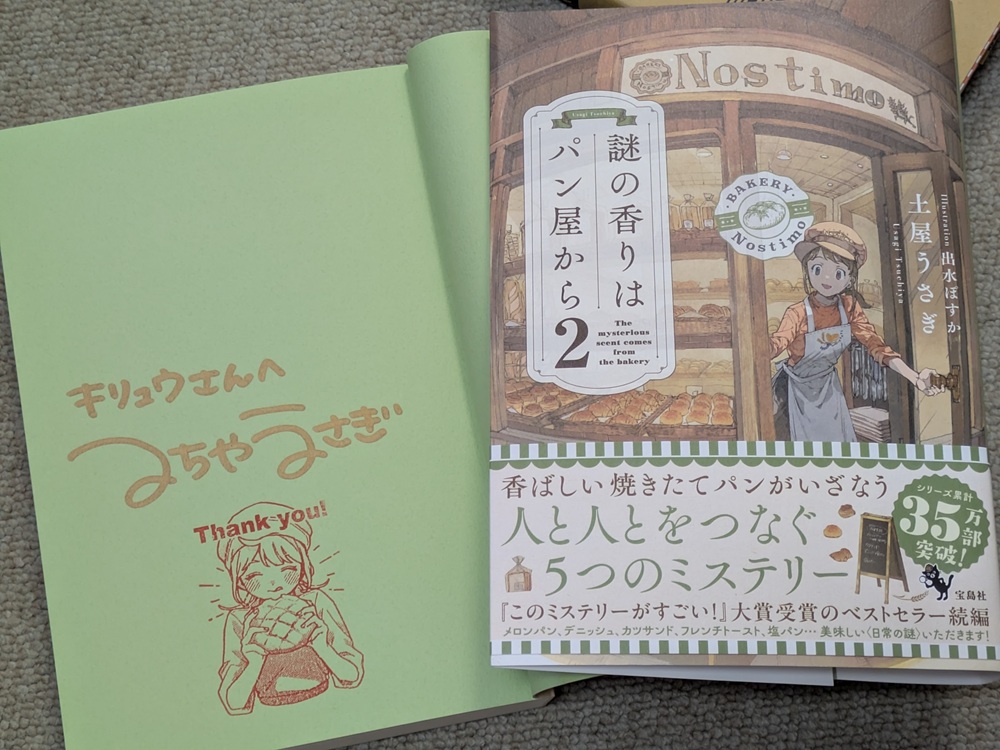 土屋うさぎ先生のサイン入り『謎の香りはパン屋から 2』(宝島社/シリーズ累計35万部突破)。グリーンの見返しに金色インクで「キリュウさんへ」のサインとメロンパンの女の子スタンプ。帯には「香ばしい焼きたてパンがいざなう 人と人をつなぐ5つのミステリー」のキャッチコピー。