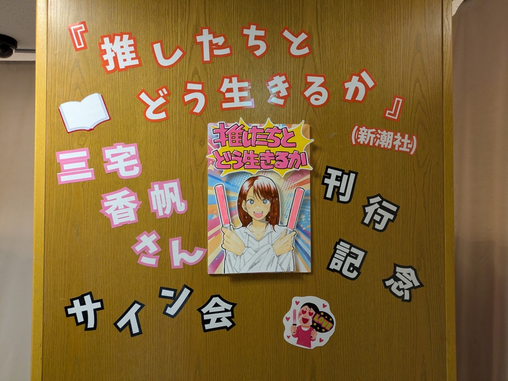 『推したちとどう生きるか』刊行記念サイン会の会場装飾。木目の壁に手描き風POPが並ぶ